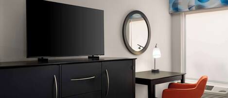 A hotel room with a flat-screen TV on a black cabinet, a round mirror on the wall, a desk with a lamp, an orange chair, and a large window with a view.
