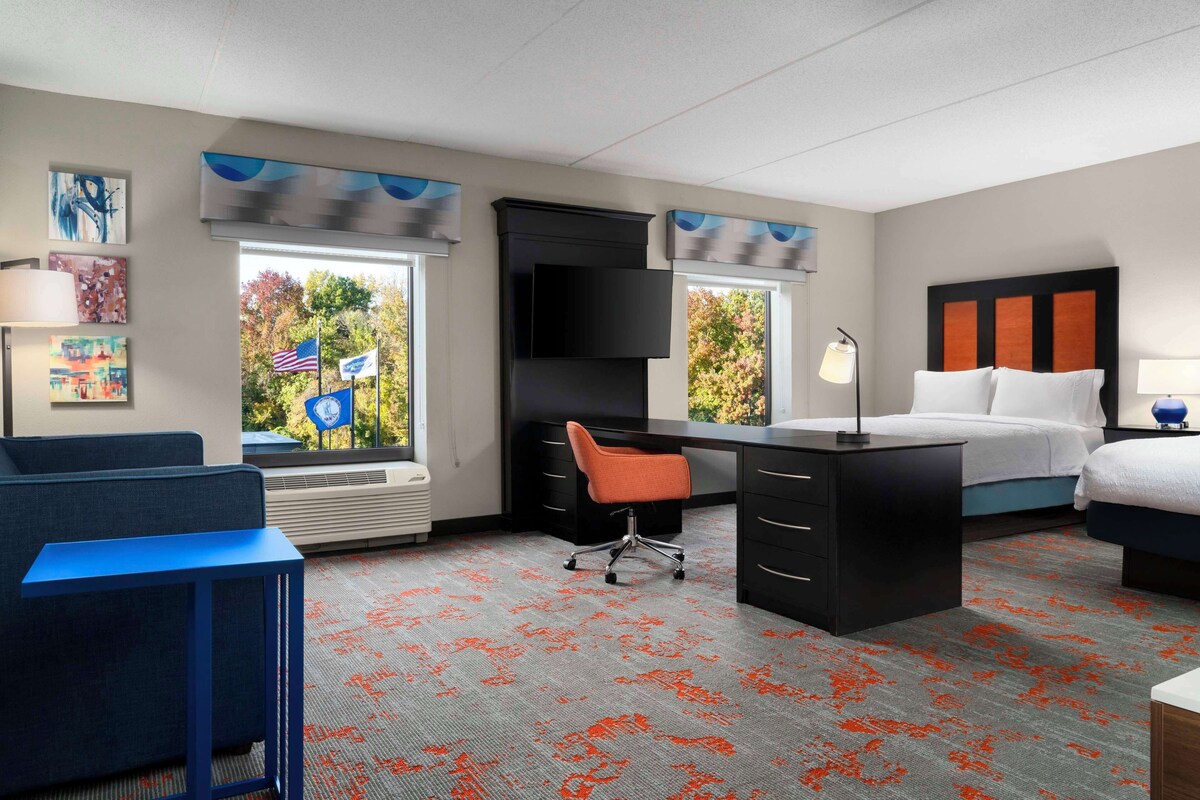 Een hotelkamer met een bureau, een bed, een stoel en uitzicht op bomen door het raam.