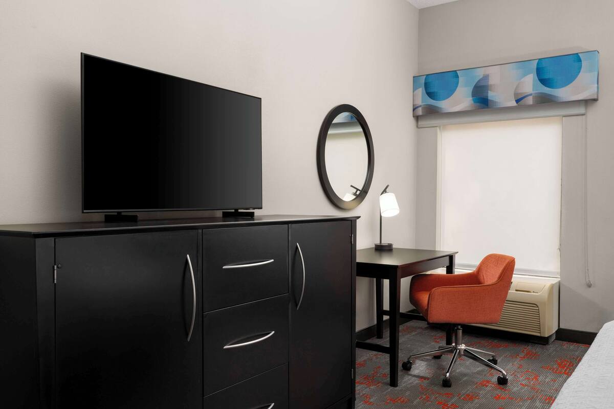 A hotel room with a TV on a cabinet, a desk with a chair, a mirror, and a window with blinds.