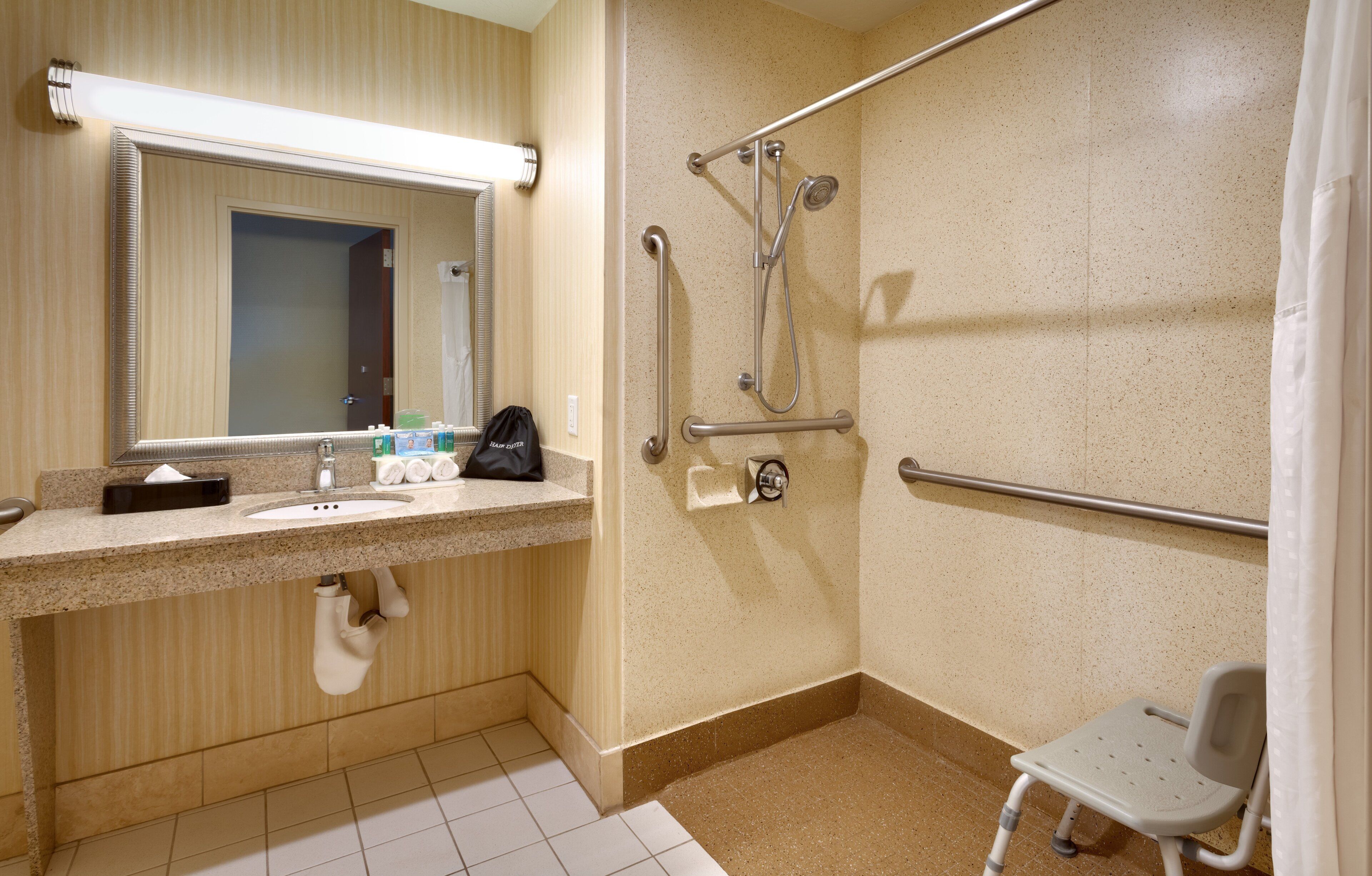 A bathroom with a sink, mirror, towel dispenser, and a shower with grab bars.