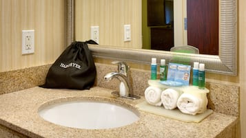 Un lavamanos de baño en hotel con un espejo, un perchero con toallas, artículos de aseo personal y una bolsa de tela negra con cordón.