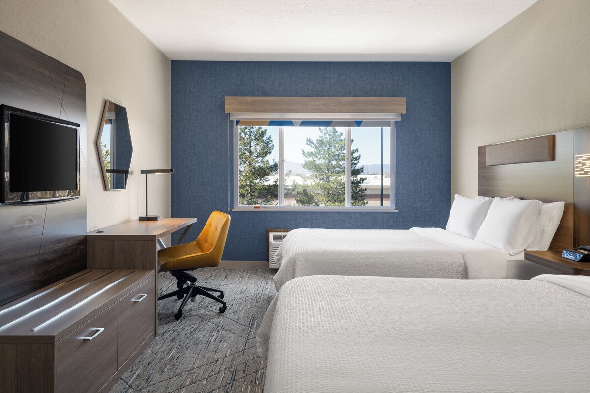 A hotel room with a bed, a desk, a chair, a TV, and a window with a view of trees and buildings.