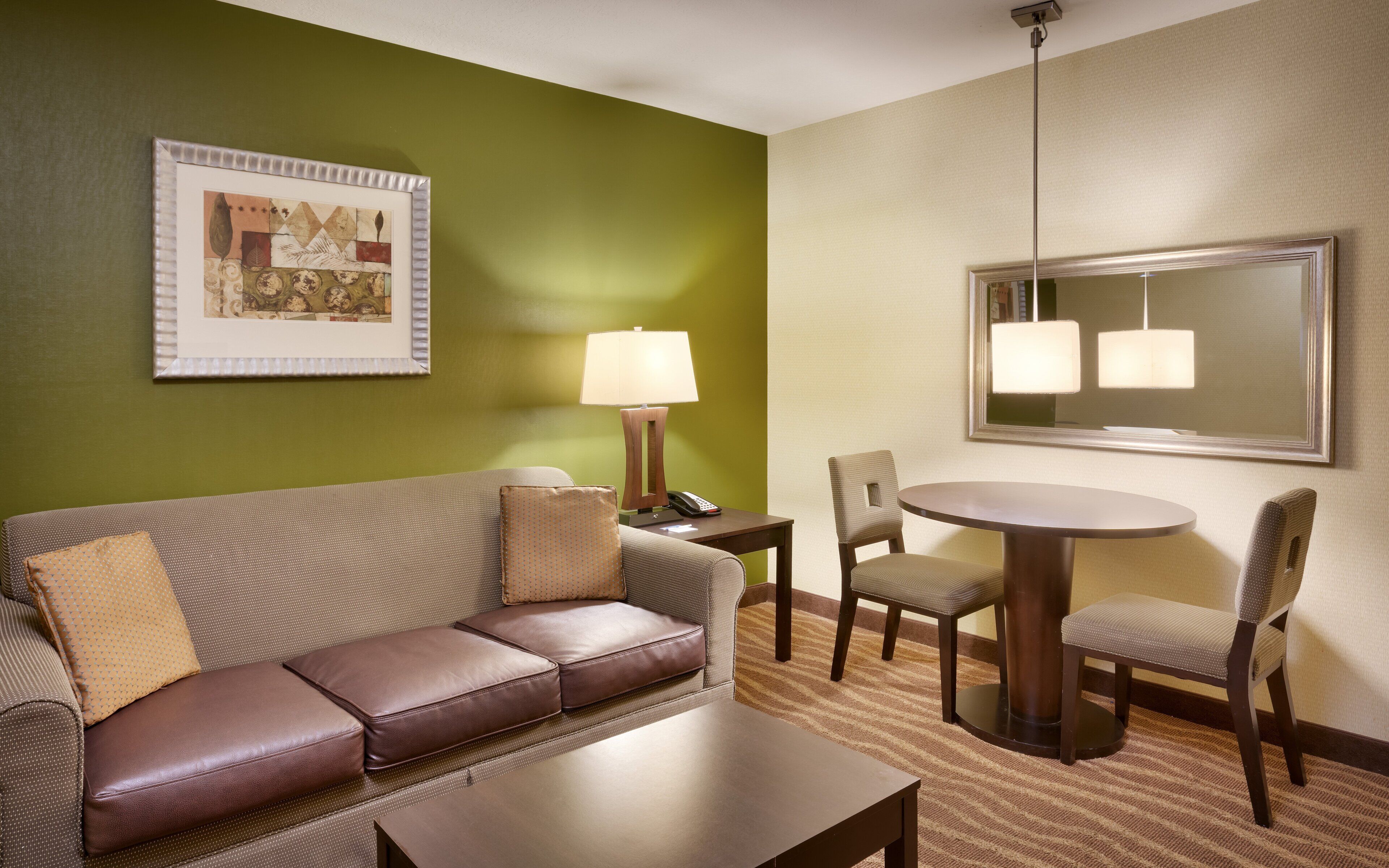 A hotel room with a green wall, a framed picture, a sofa with leather cushions, a small table, a lamp, a round dining table with chairs, and a mirror above the dining table.