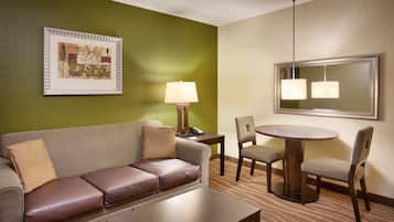 A hotel room with a green wall, a framed picture, a sofa with leather cushions, a small table, a lamp, a round dining table with chairs, and a mirror above the dining table.