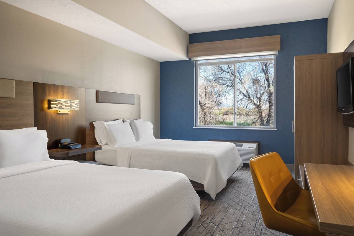A hotel room with two beds, a wooden desk, a television, and a window with a view of trees.