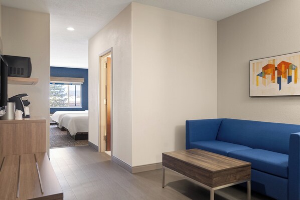 A modern hotel room with a blue sofa, a wooden coffee table, a TV, and a bed with white bedding. - Holiday Inn Express & Suites American Fork - North Provo by IHG (American Fork)