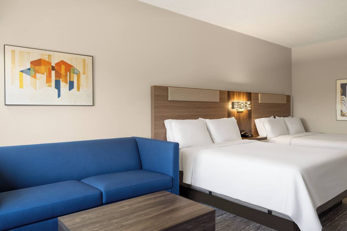 A hotel room with a blue sofa, two beds, a wooden headboard, and a wall-mounted light fixture.
