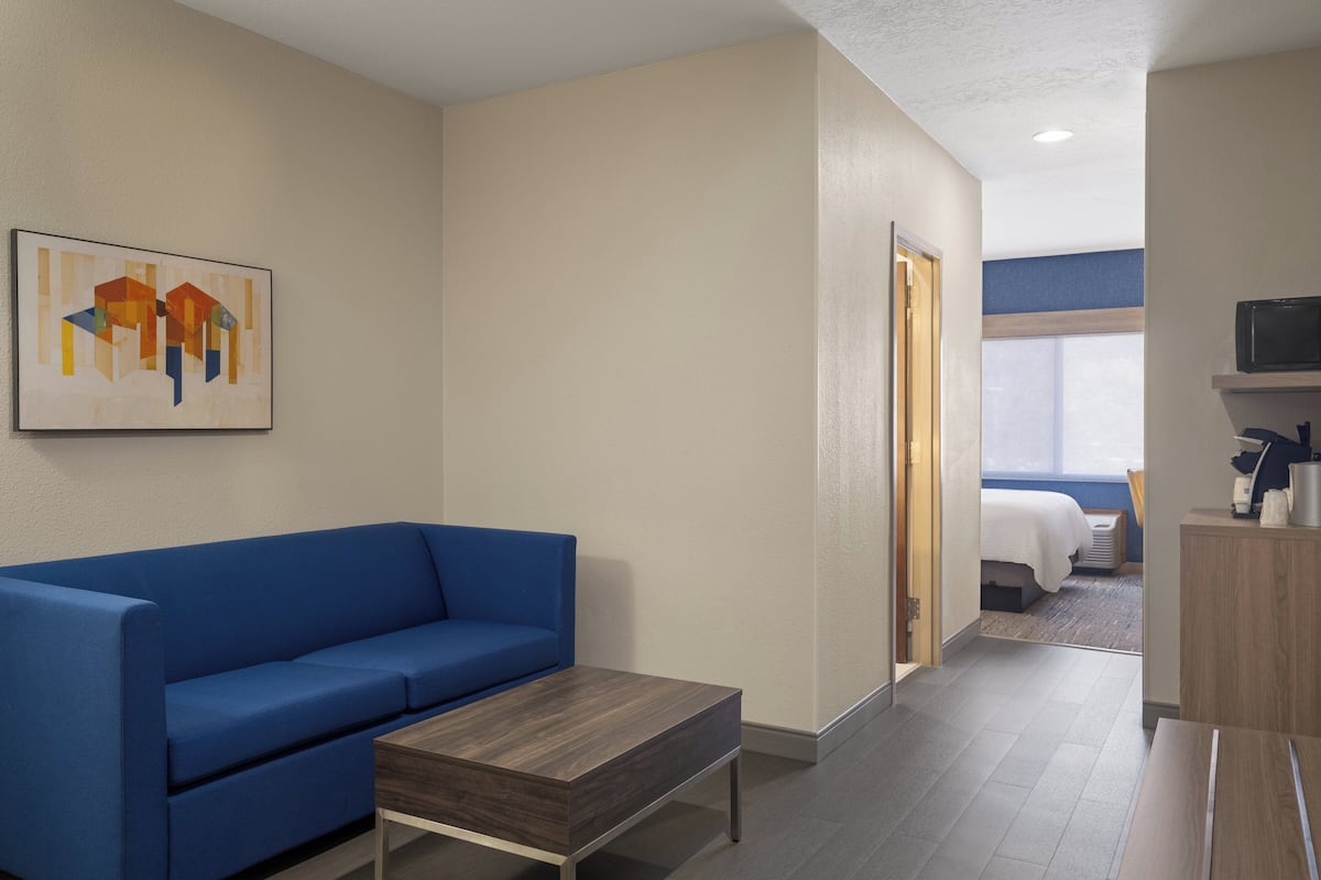 A hotel room with a blue sofa, a wooden coffee table, a painting on the wall, a television on a wooden stand, and a bed with a white comforter.