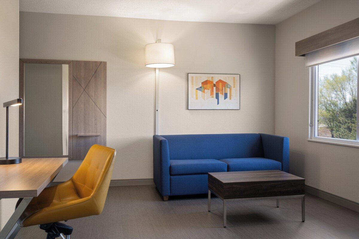 A modern living room with a blue sofa, a wooden coffee table, a yellow chair, a floor lamp, and a framed abstract artwork on the wall.