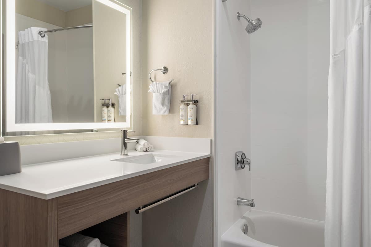 A modern bathroom with a white sink, mirror, and bathtub. There are towels and toiletries on the counter.