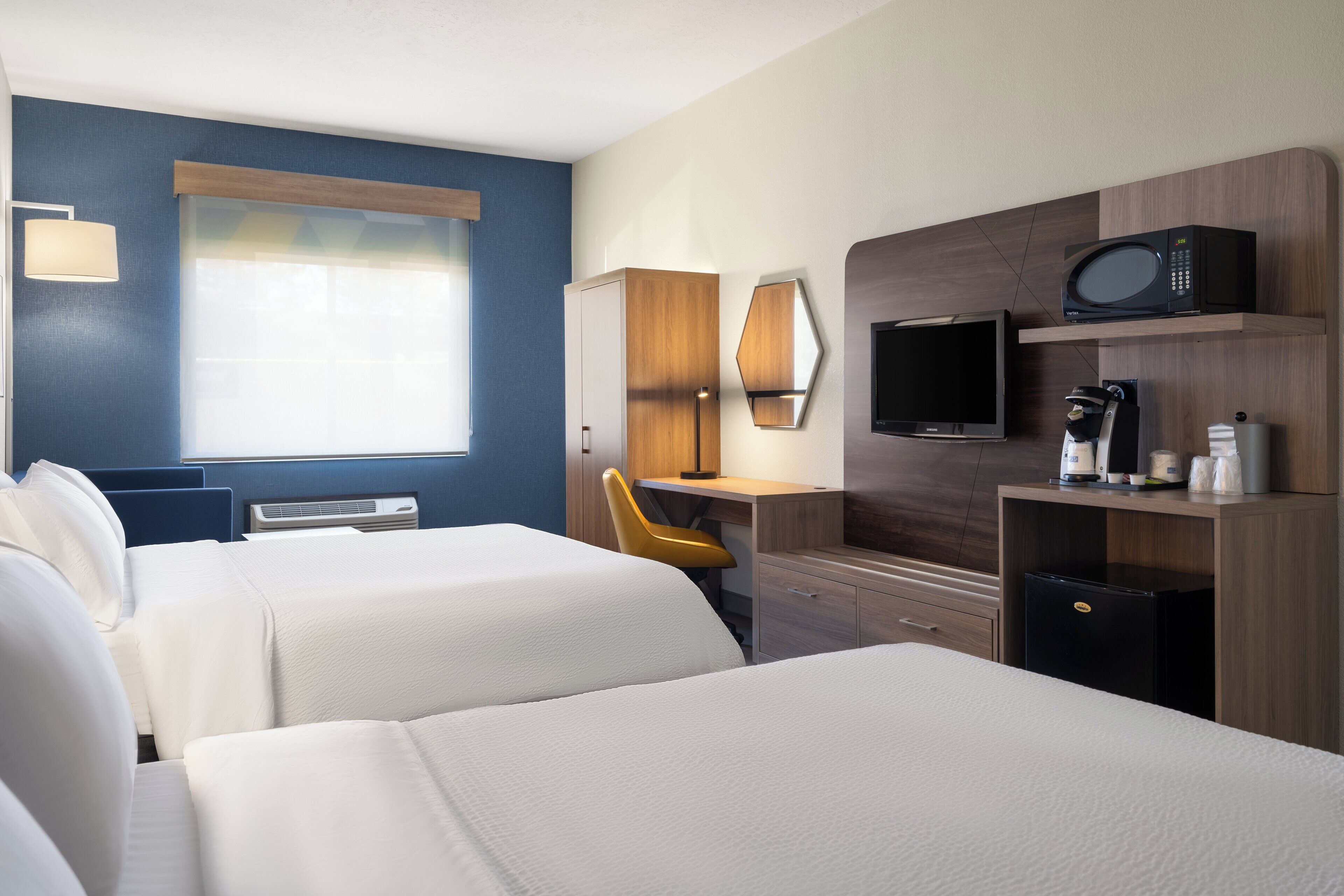 A hotel room with two beds, a desk, a chair, a TV, a coffee maker, and a refrigerator.