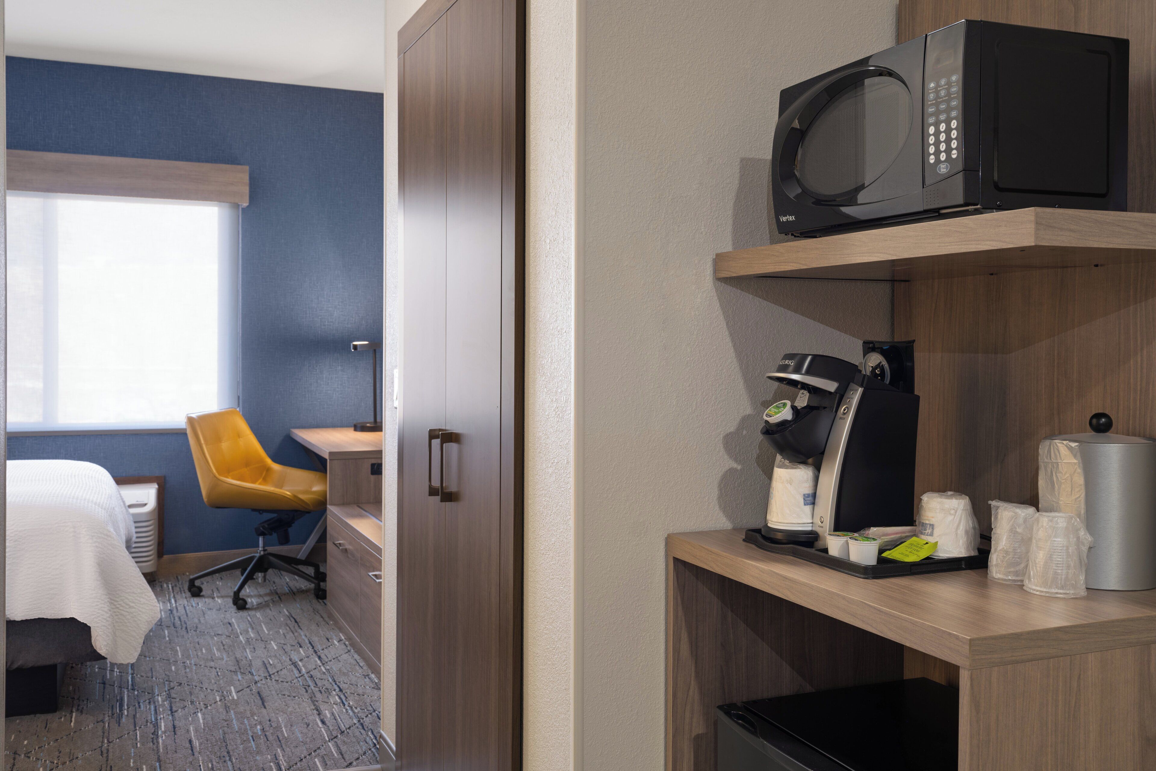 Ein Hotelzimmer mit Bett, Schreibtisch, Stuhl, einer Mikrowelle, einer Kaffeemaschine und einem Regal mit Pflegeprodukten.