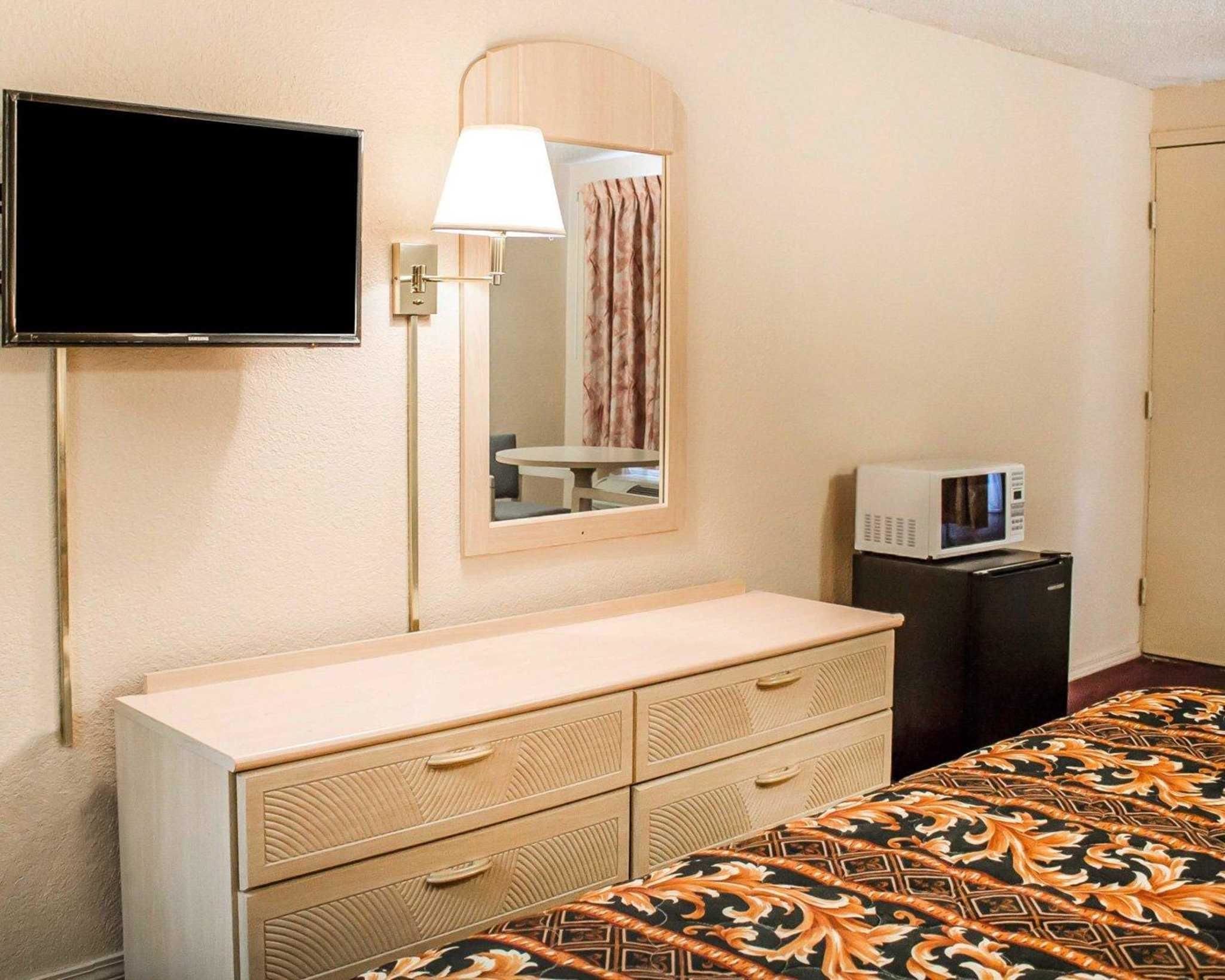 A hotel room with a bed, a dresser with drawers, a television mounted on the wall, a lamp, a mirror, a microwave, and a curtain.