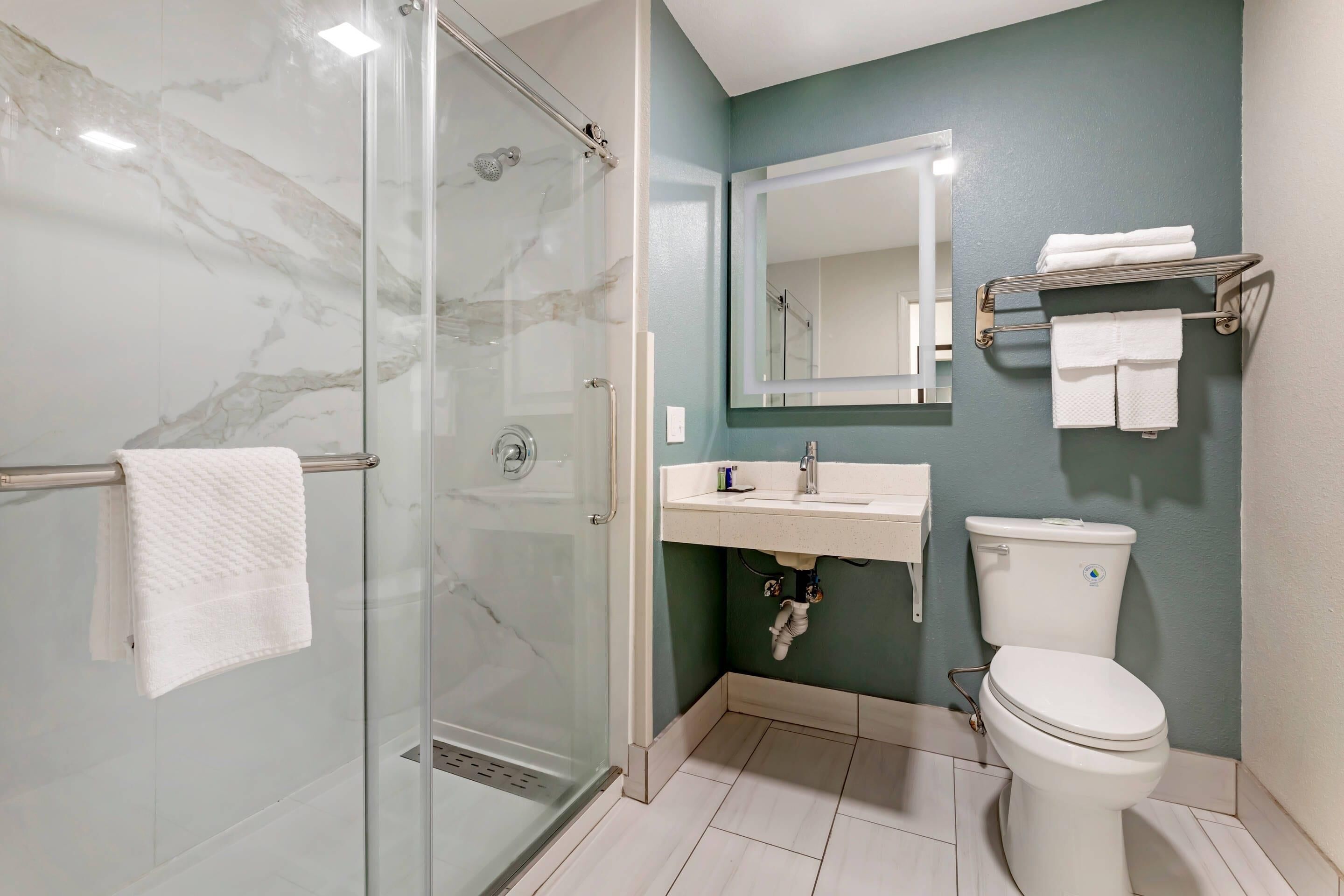 A bathroom with a glass shower enclosure, a white toilet, a sink with a mirror above it, and a towel rack with folded towels.