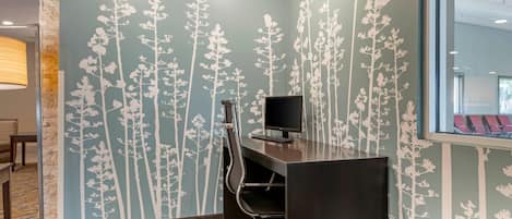 Un espacio de oficina moderno con un escritorio, una silla y una computadora. Las paredes tienen un estampado floral y hay una ventana con sillas rojas a la vista.