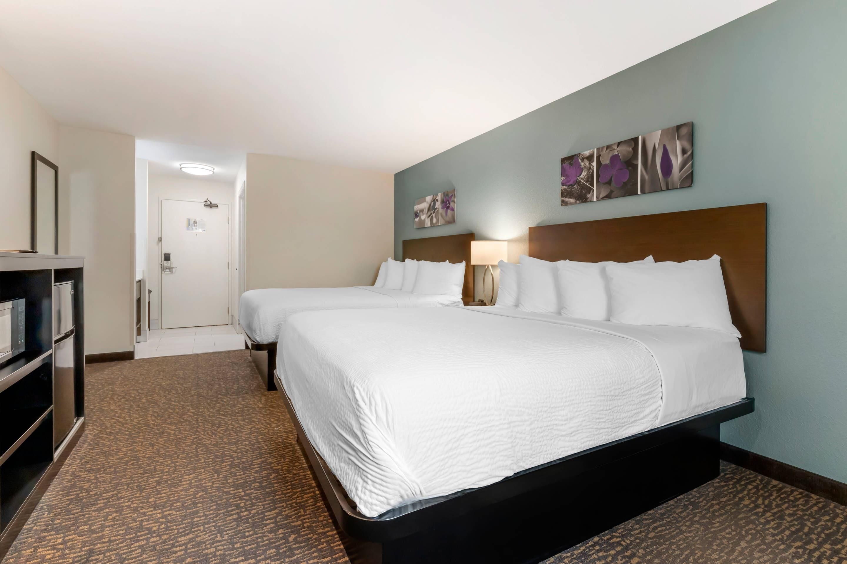 Habitación de hotel con dos camas, una mesita de noche con lámpara, un armario y una puerta que da a un baño.