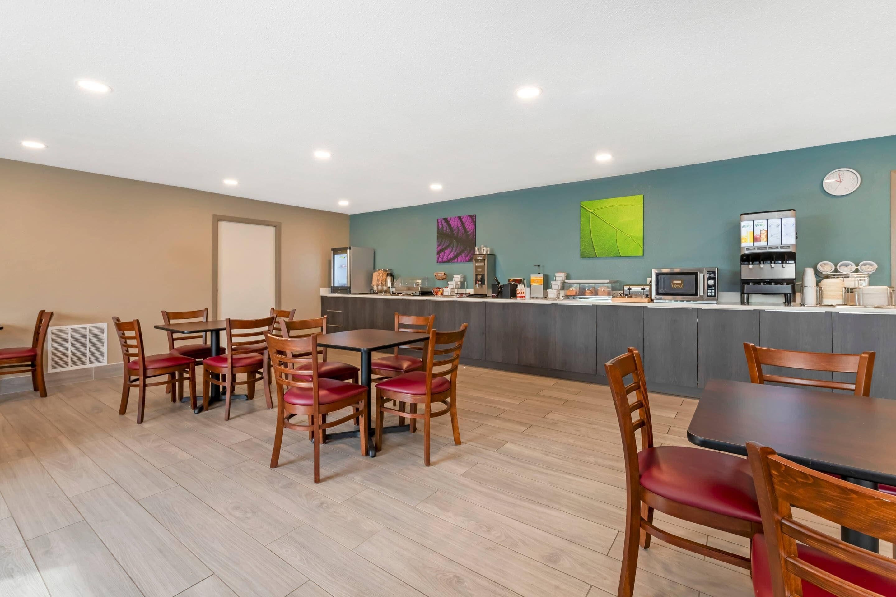 A modern dining area with wooden tables and chairs, a kitchen counter with coffee machines, and a green wall with abstract art.