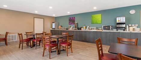 A modern dining area with wooden tables and chairs, a kitchen counter with coffee machines, and a green wall with abstract art.