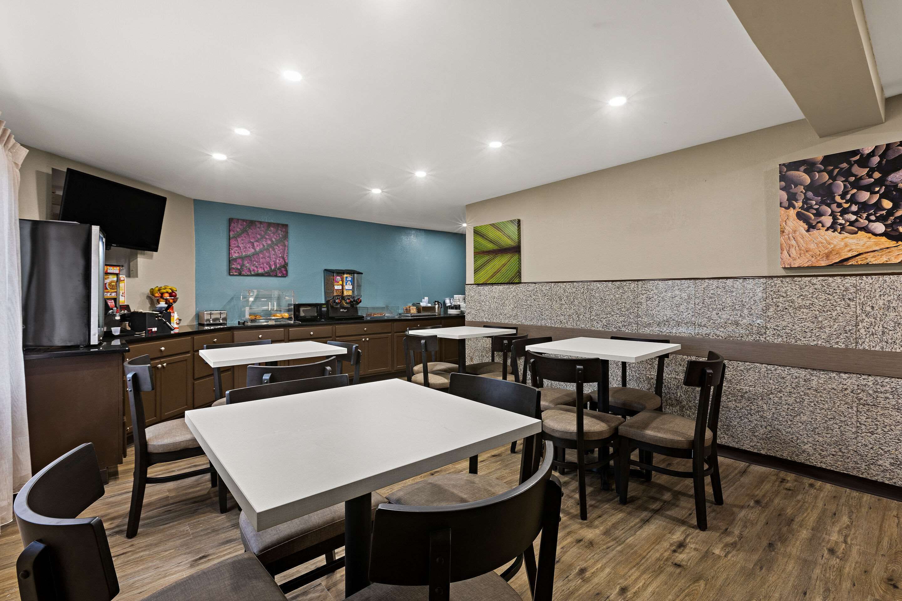 A modern restaurant with multiple tables and chairs, a kitchen area, and a flat-screen TV mounted on the wall.