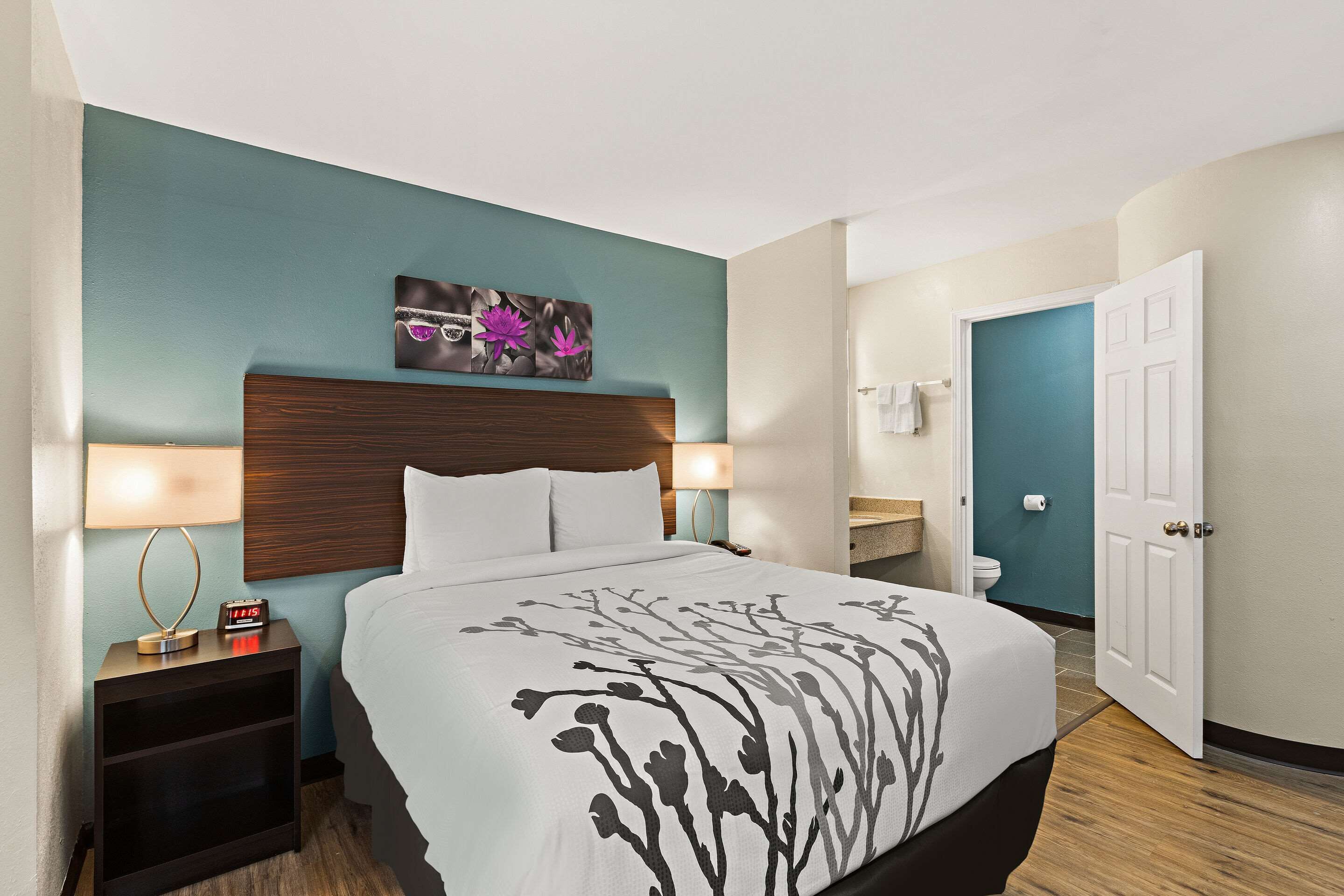 A hotel room with a bed, bedside tables, a nightstand, a wall-mounted clock, a painting, a bathroom with a toilet, and a door.