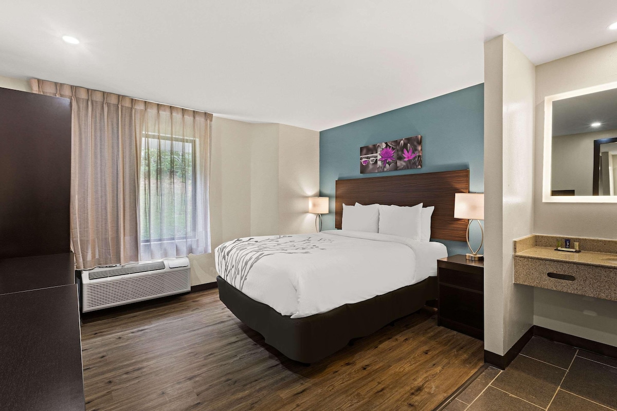 A hotel room with a bed, bedside tables, a mirror, a window with curtains, and a wall-mounted artwork.