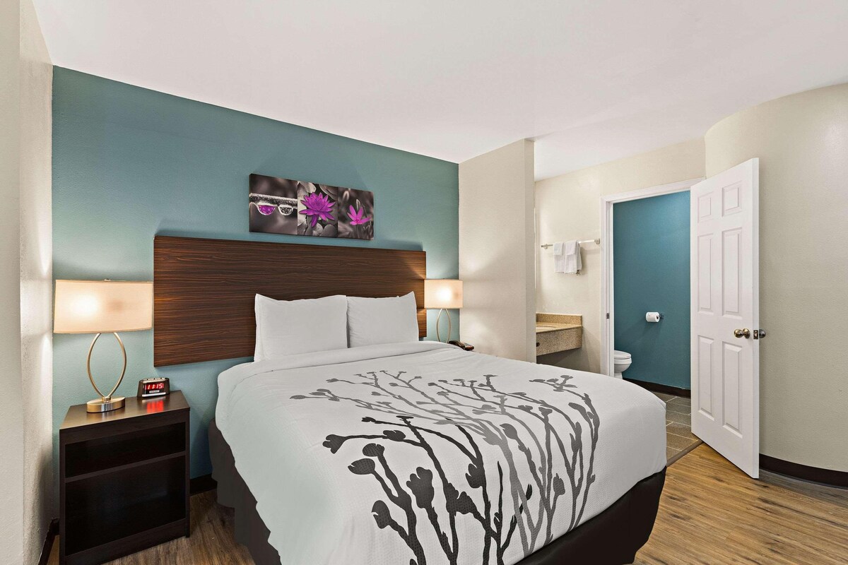 A hotel room with a bed, bedside tables, a nightstand, a wall-mounted clock, a bathroom with a toilet and sink, and a door to the bathroom.