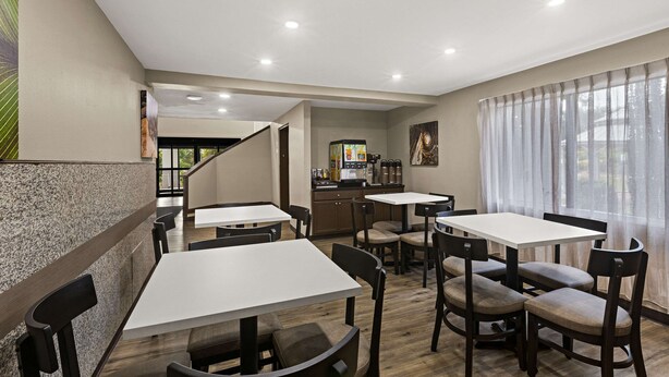 Un comedor moderno con varias mesas y sillas, una cocina al fondo y una escalera que sube a otro piso.