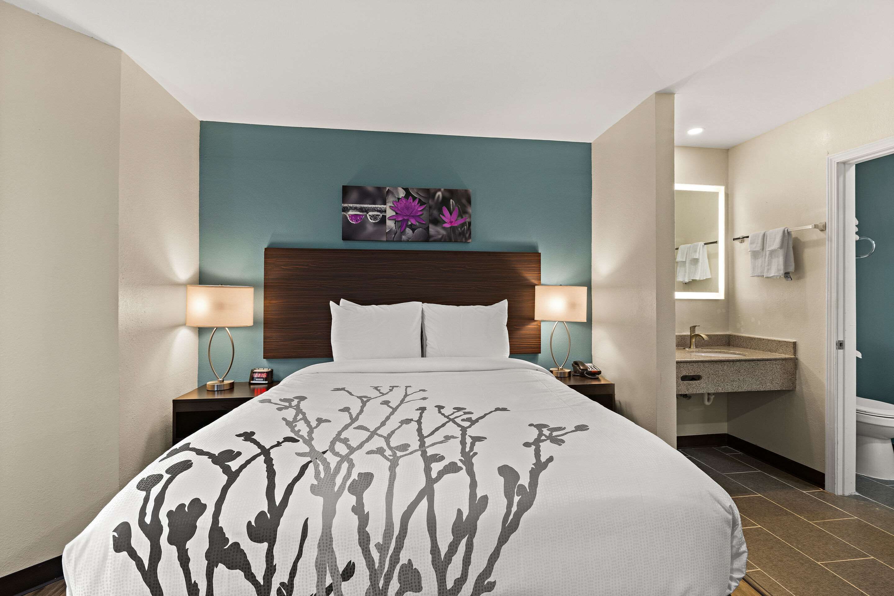 Una habitación de hotel con una cama, lámparas de noche, una mesita de noche, un baño con lavamanos y espejo, y una obra de arte en la pared sobre la cama.