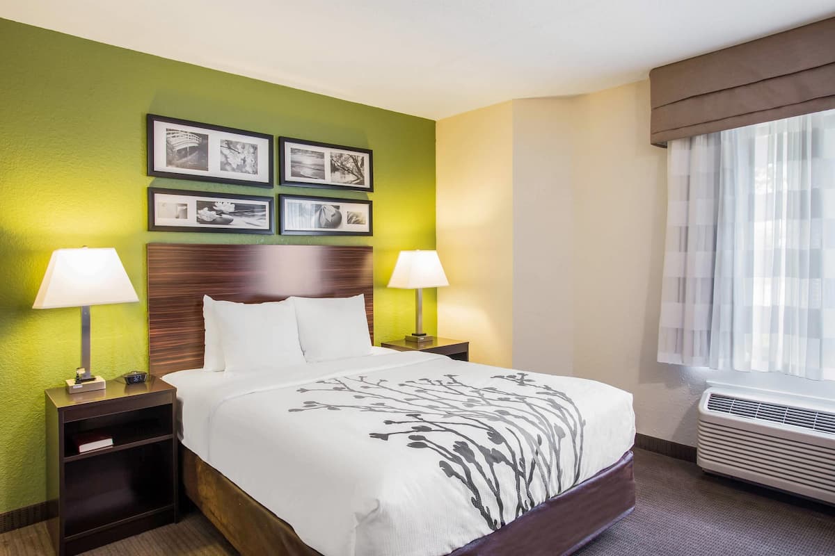 Habitación de hotel con cama, dos mesitas de noche con lámparas, un aparador y una ventana con cortinas. En la pared, varias fotografías enmarcadas.