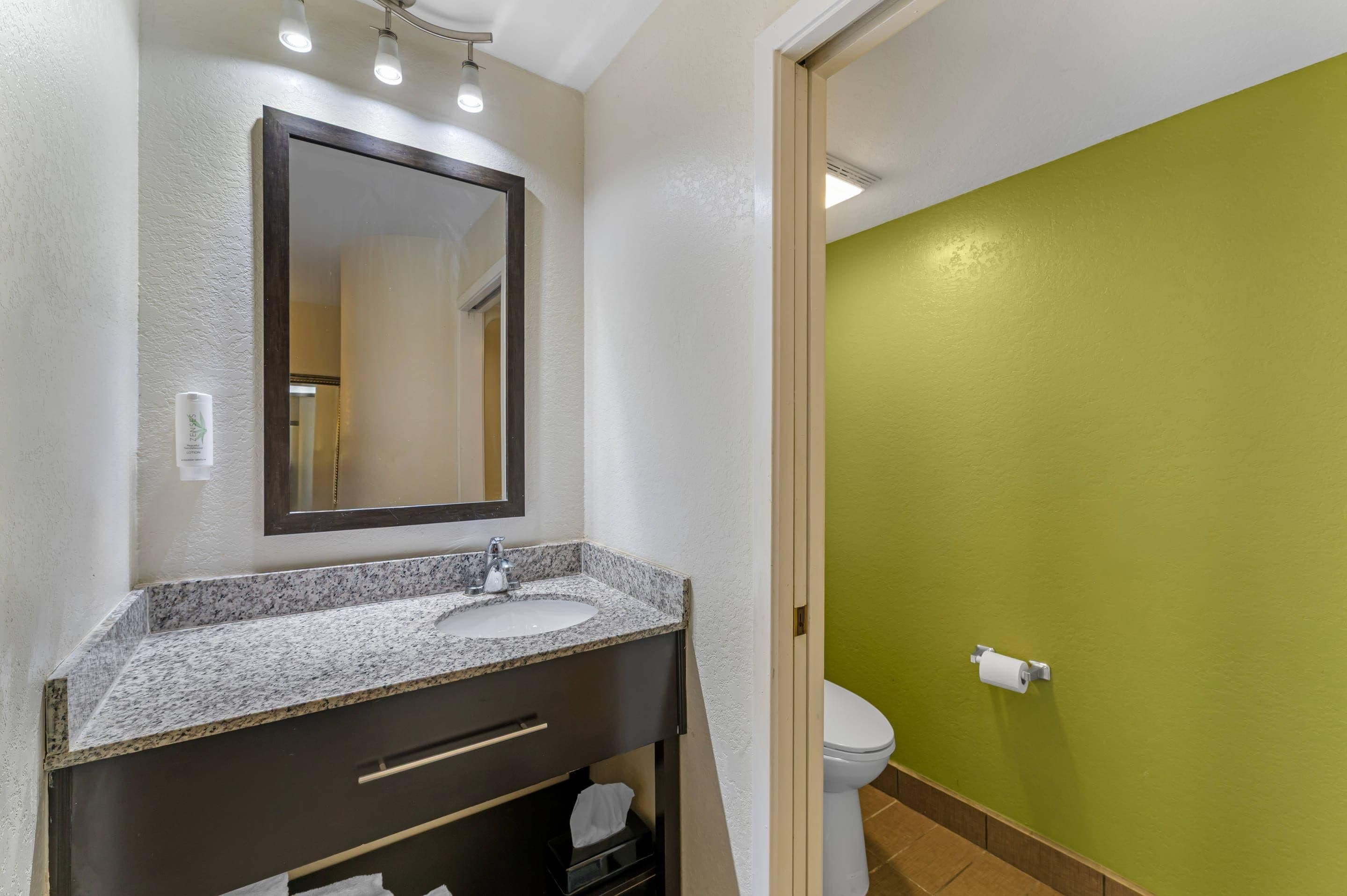 A bathroom with a granite countertop sink, a dark vanity, a mirror, and a toilet against a green wall.