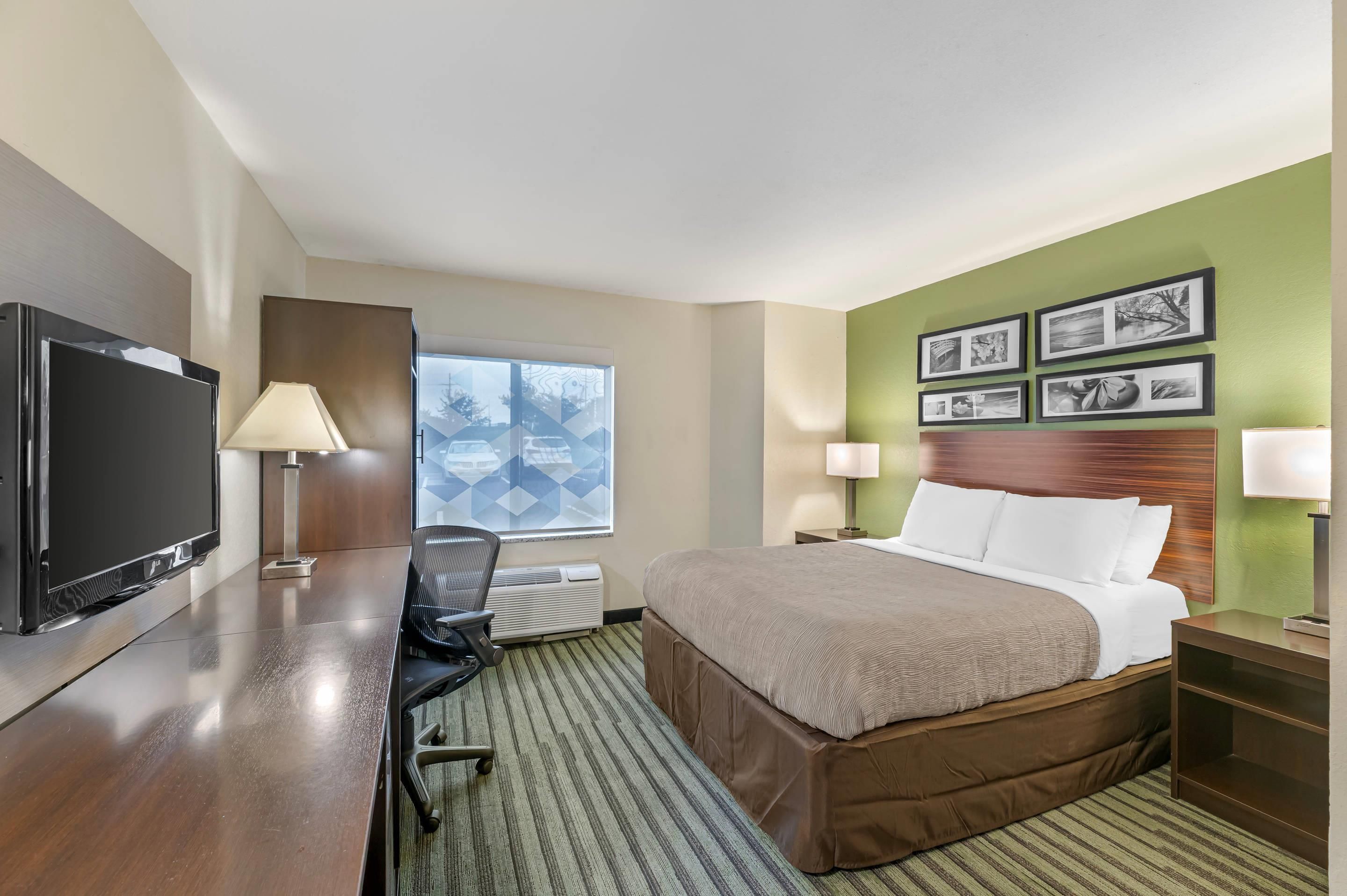 Habitación de hotel con cama, escritorio con televisor, silla y ventana con vista a edificios.