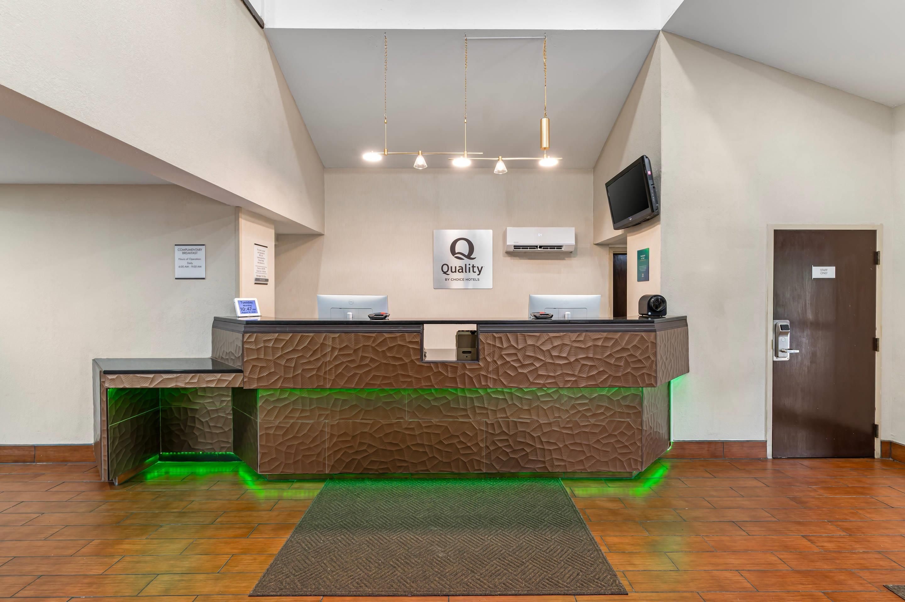 A hotel reception desk with a 'Quality by Choice Hotels' sign, a flat-screen TV, and a digital clock.