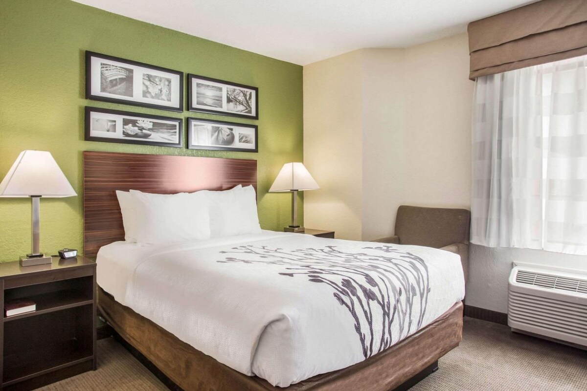 Ein Hotelzimmer mit einem Bett, zwei Nachttischen, einem Wandbild und einem Fenster mit Vorhängen sowie einer Klimaanlage.