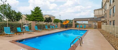 A rectangular outdoor swimming pool with a brick border, surrounded by a white fence and blue lounge chairs.