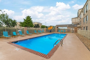 A rectangular outdoor swimming pool with a brick border, surrounded by a white fence and blue lounge chairs.