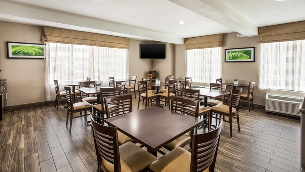 A hotel dining area with multiple tables and chairs, a flat-screen TV mounted on the wall, and large windows with curtains.