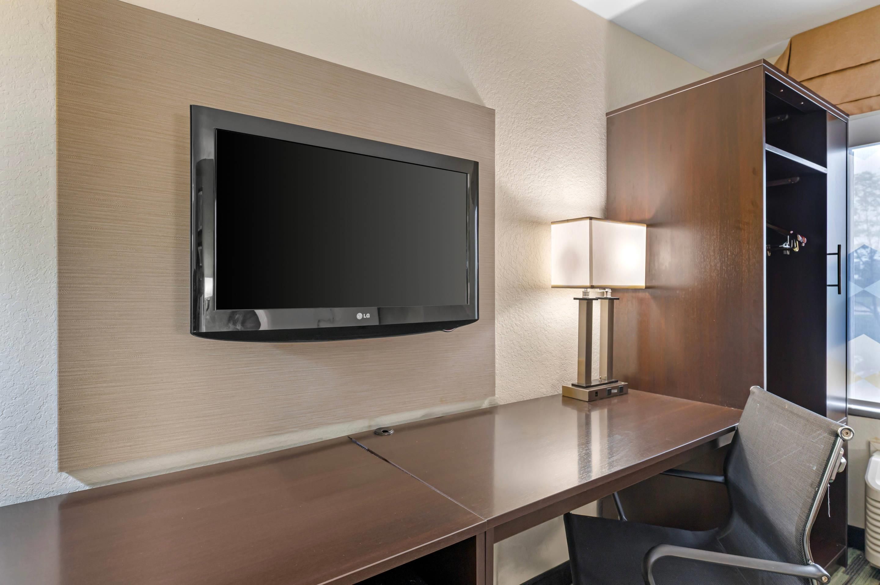 A hotel room with a flat-screen TV mounted on the wall, a desk with a chair, and a lamp.
