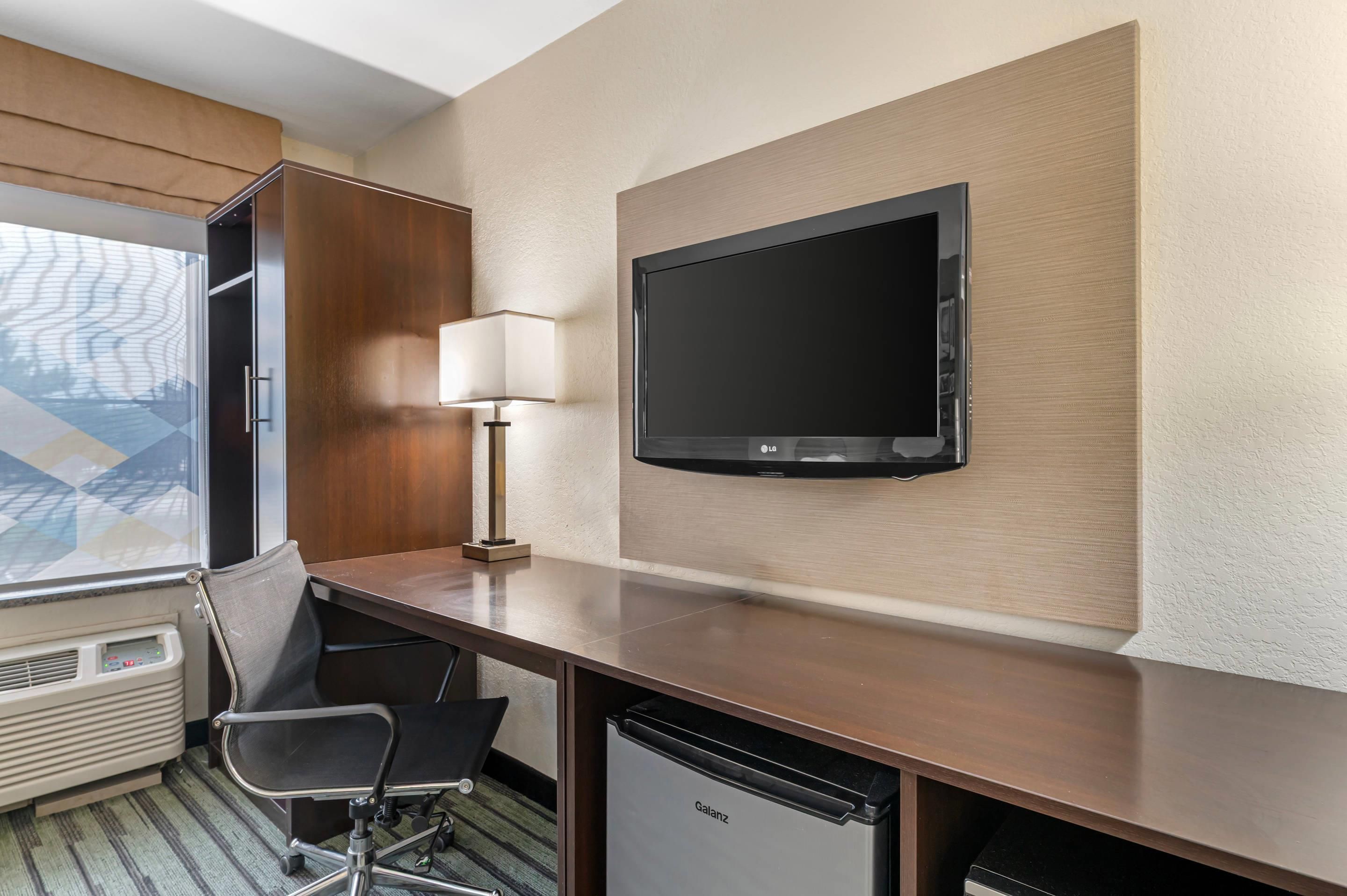 Habitación de hotel con escritorio, una silla, televisor montado en la pared y ventana con vista a un edificio.
