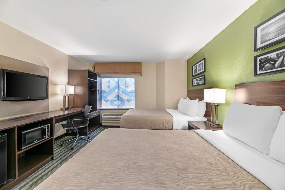 Une chambre d’hôtel comprenant un lit, un bureau avec un téléviseur à écran plat, une chaise, une lampe de bureau et une fenêtre ornée d’un motif géométrique.
