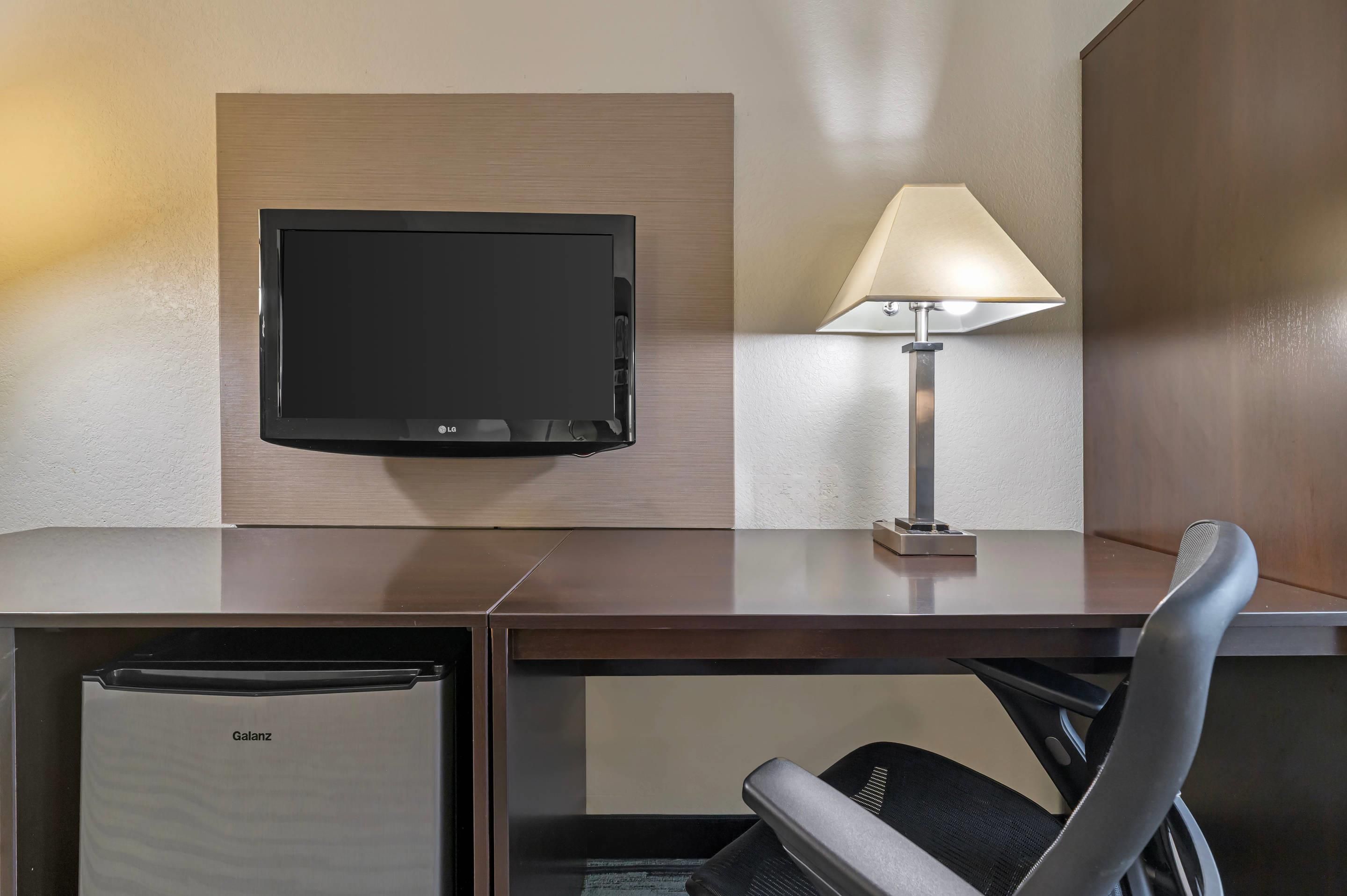 Habitación de hotel con televisor de pantalla plana montado en la pared, escritorio con lámpara, silla y minibar.