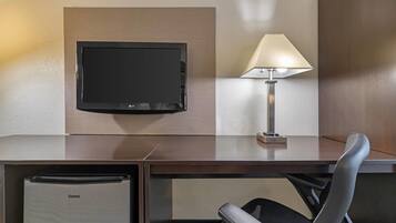 Habitación de hotel con televisor de pantalla plana montado en la pared, escritorio con lámpara, silla y minibar.