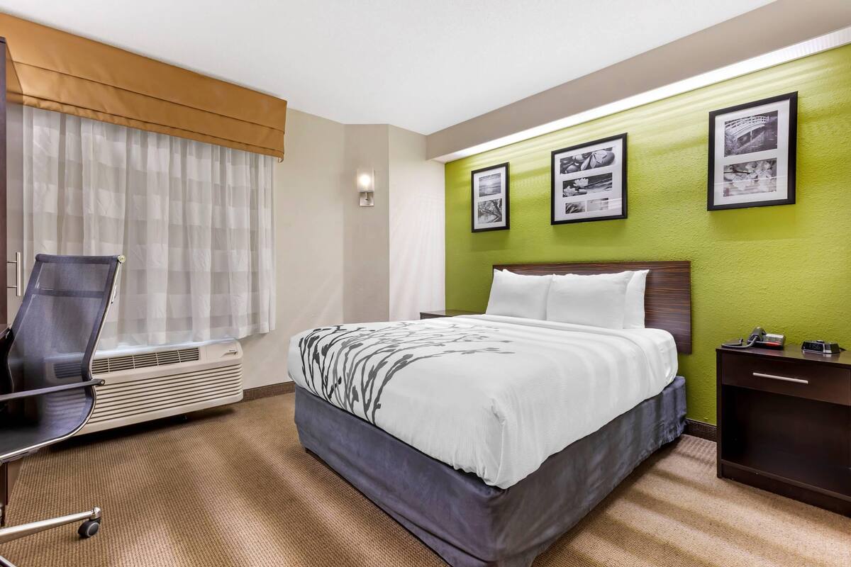 A hotel room with a bed, a desk, a chair, an air conditioner, and a nightstand.