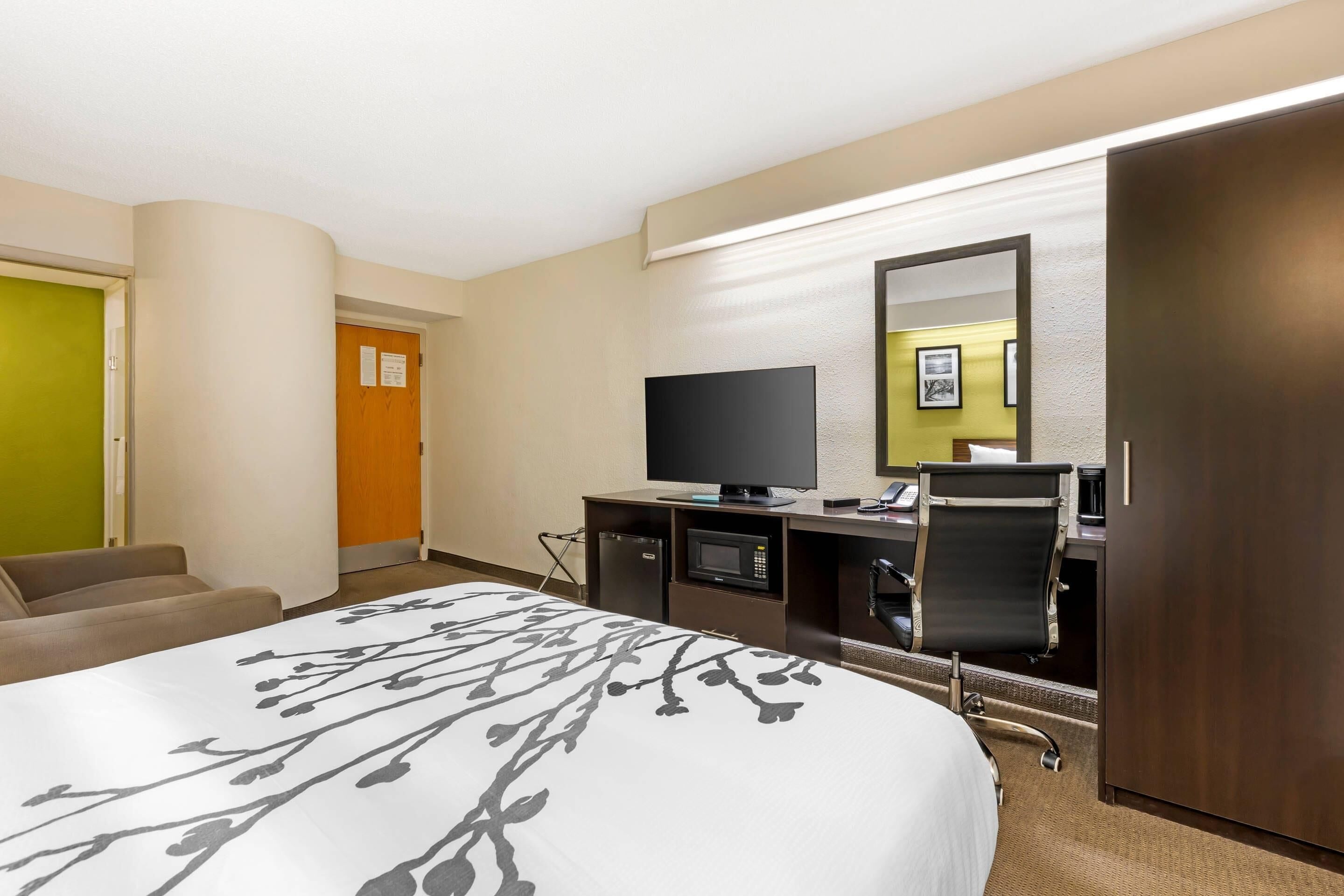Una habitación de hotel con una cama, televisión, escritorio con computadora, silla y un refrigerador pequeño.