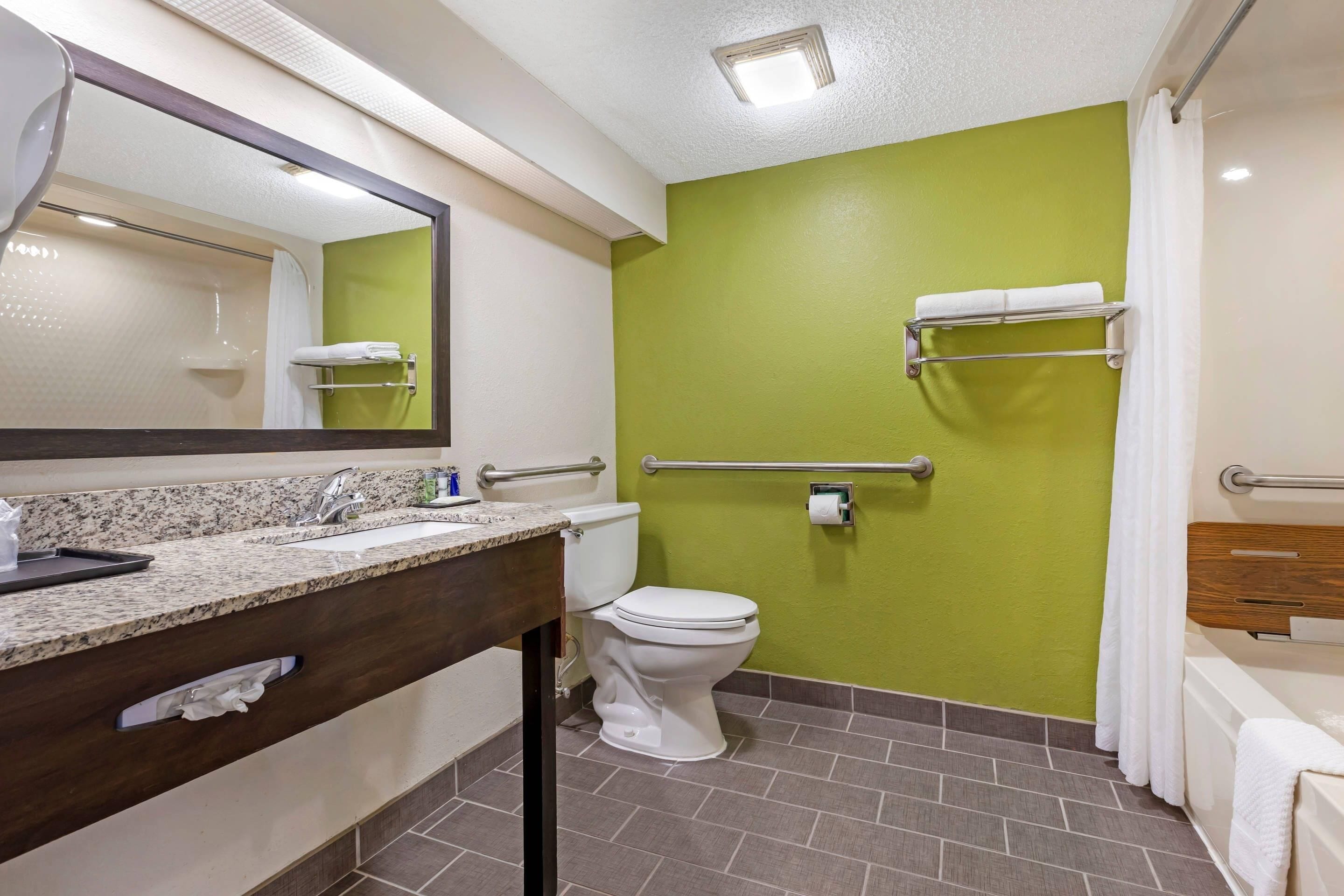 Un baño con pared verde, inodoro blanco, lavamanos con espejo y zona de ducha.