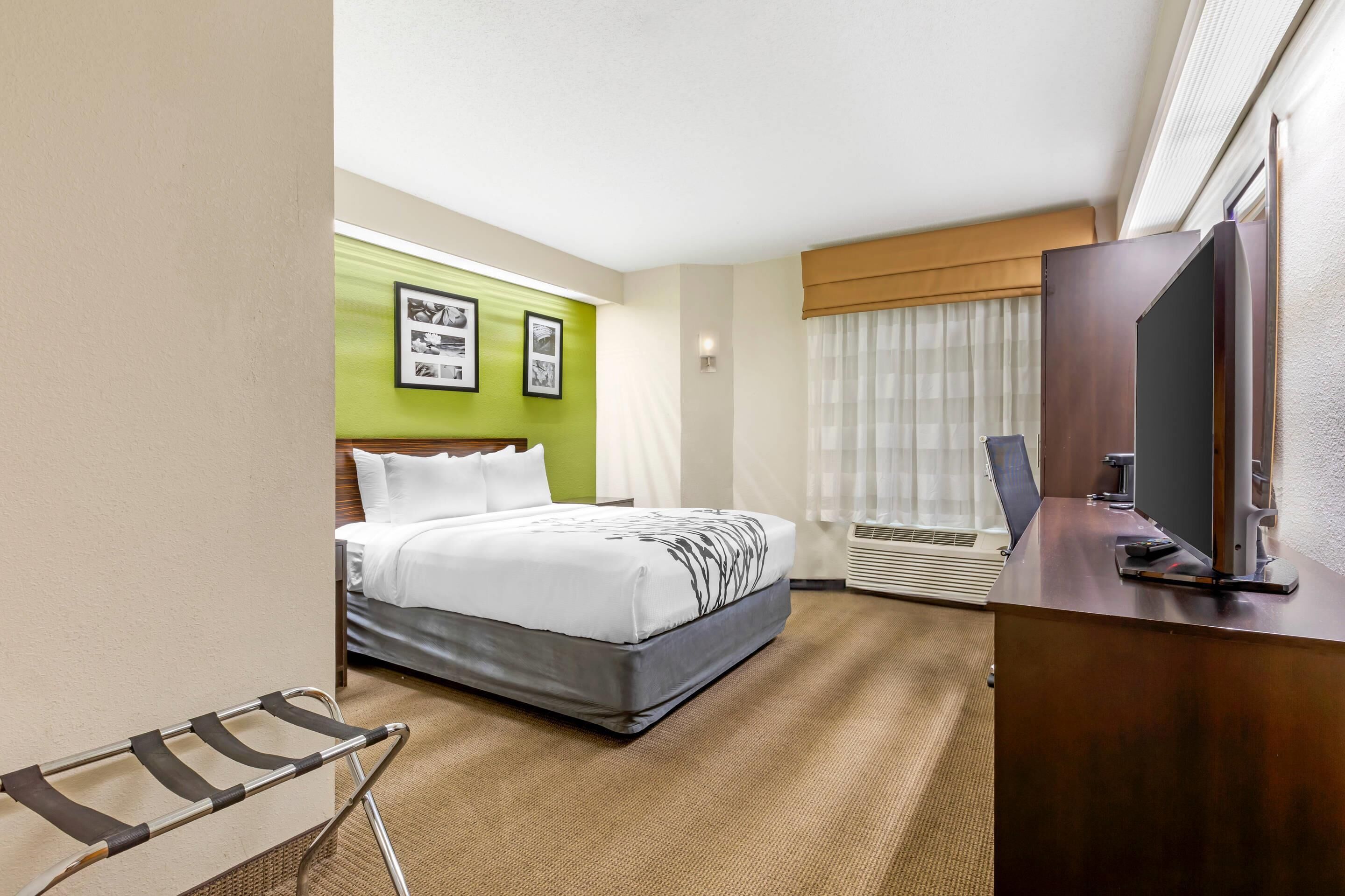 Una habitación de hotel con cama, escritorio con televisor, ventana con cortinas y aire acondicionado de pared.