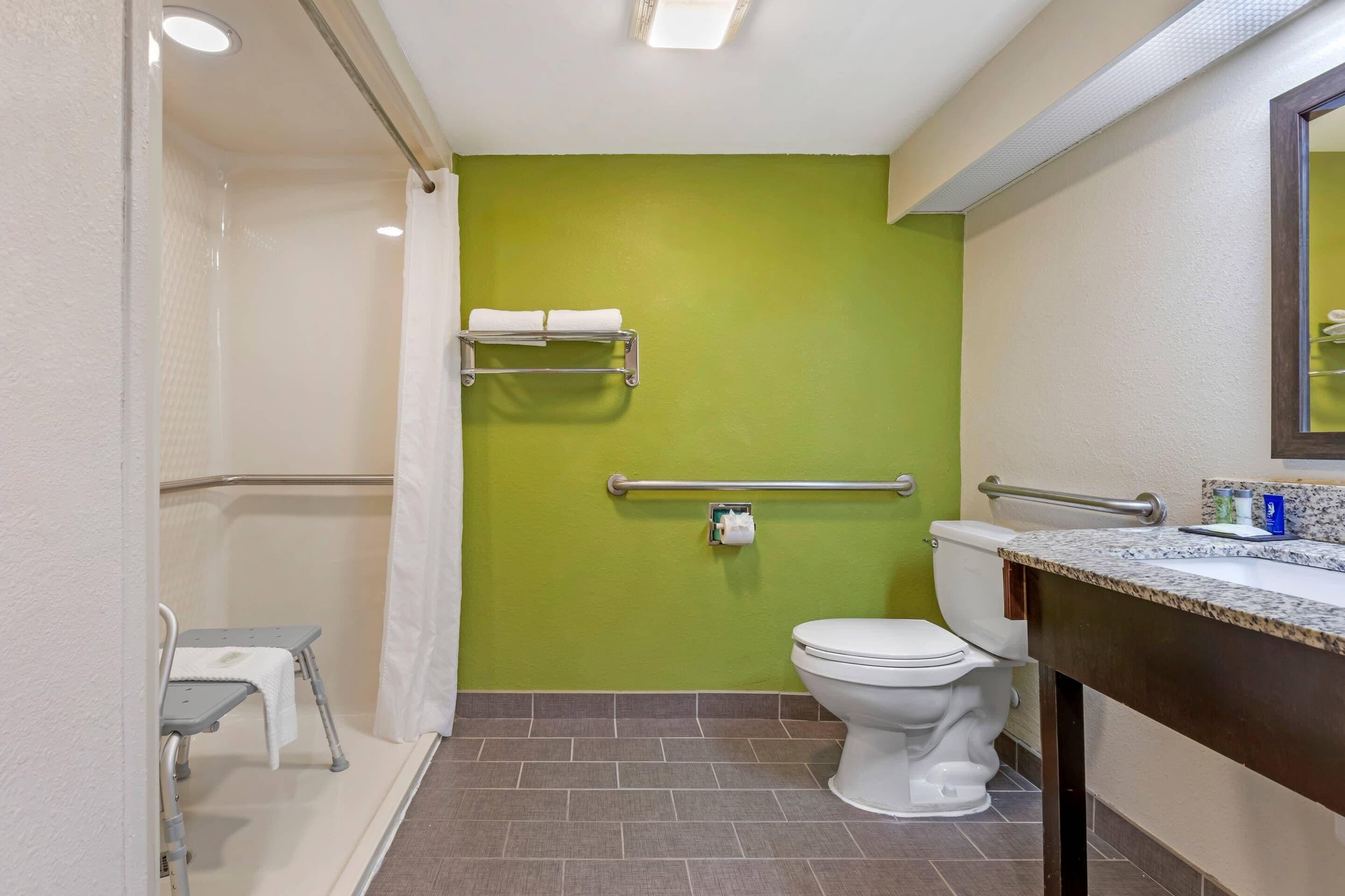 A bathroom with a green wall, a white toilet, a shower with a grab bar, and a small bench.