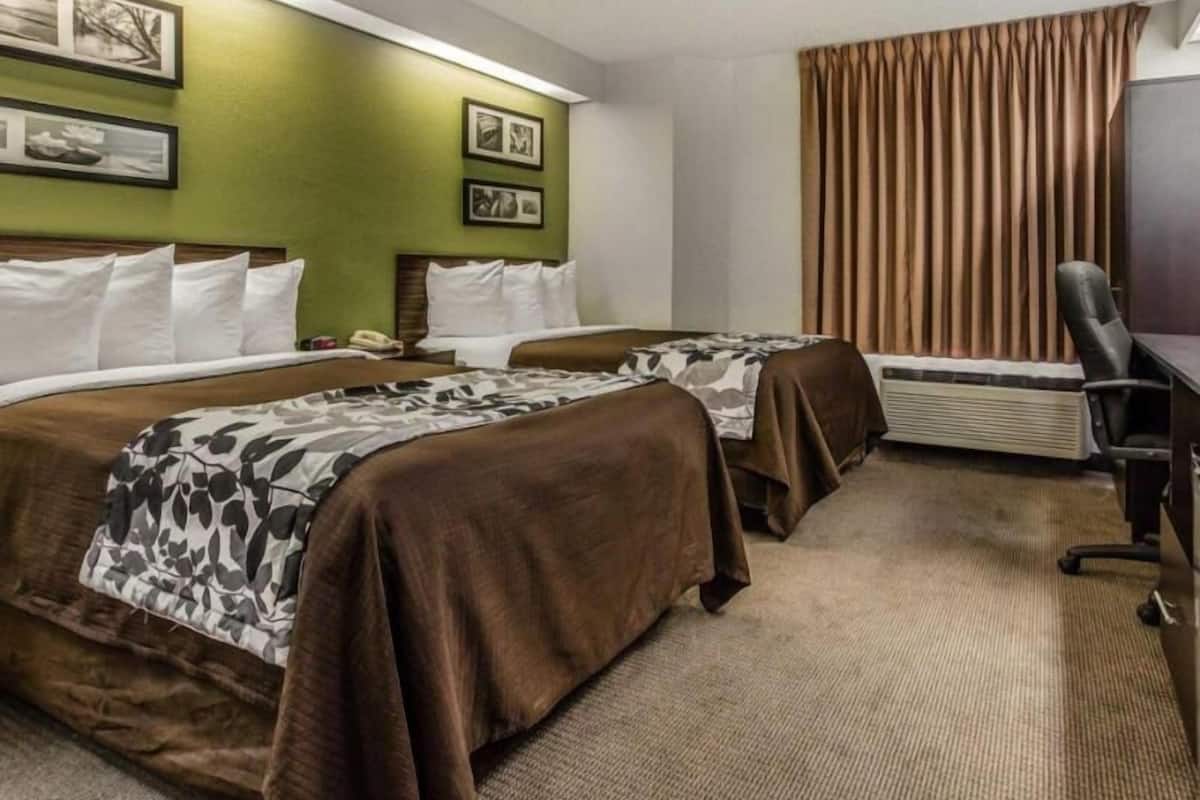 A hotel room with two beds, a desk, an air conditioner, and framed pictures on the wall.
