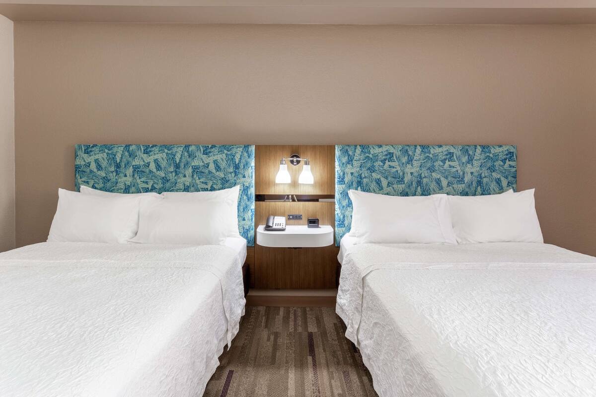 Une chambre d’hôtel avec deux lits, une tête de lit en bois, une table de chevet avec une lampe et un petit lavabo.