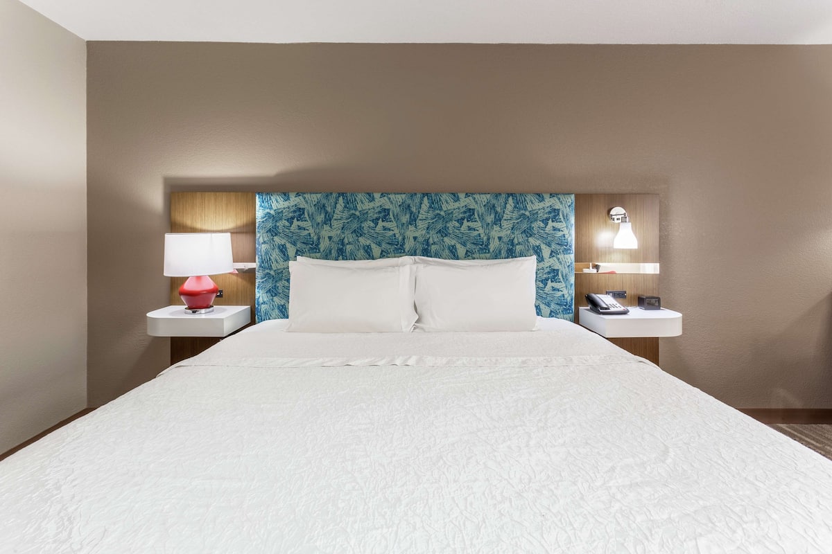 Une chambre d’hôtel avec un grand lit, deux tables de chevet, une petite table, une lampe rouge, une lampe blanche et une tête de lit bleue à motifs.