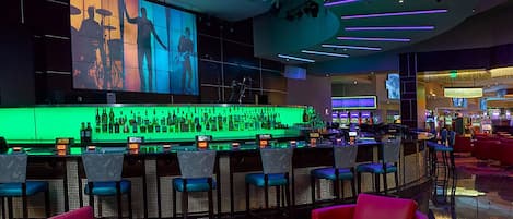 Eine Bar mit beleuchteter Theke in Grün, verschiedenen Sitzgelegenheiten und einem großen Bildschirm, der Silhouetten von Personen zeigt.